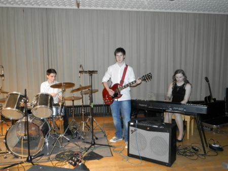 SchÃ¼lergruppe macht Musik. Ein SchÃ¼ler spielt Schlagzeug, ein weiterer Gitarre, eine SchÃ¼lerin spielt Keyboard. Im Vordergrund steht ein GitarrenverstÃ¤rker.