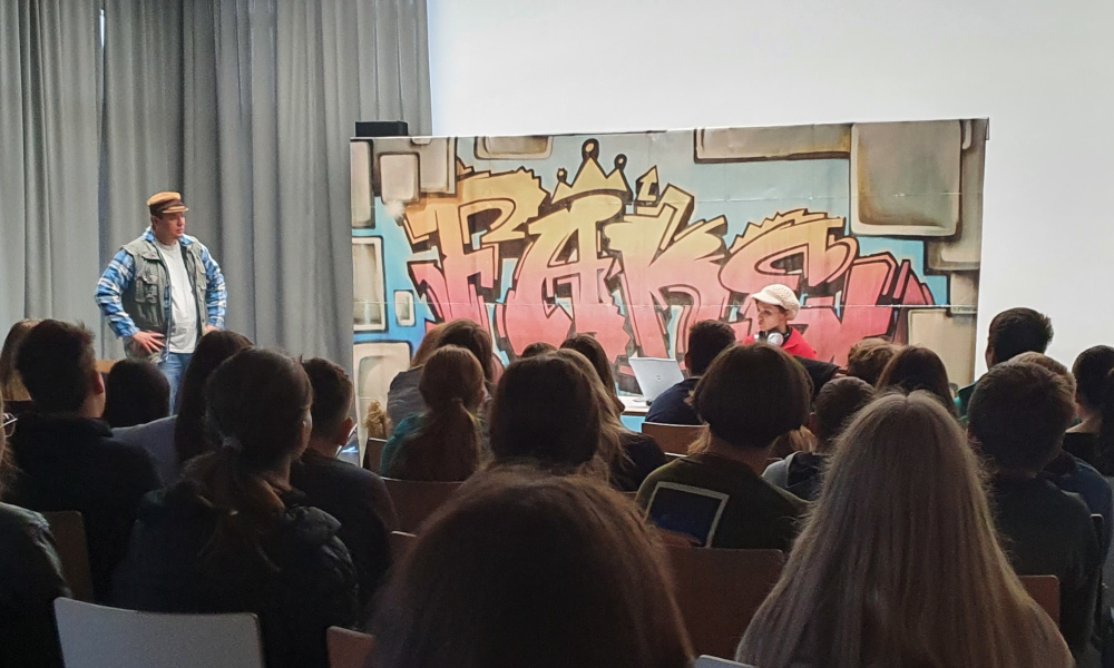 Szene aus dem Theaterstück "Fake". Schüler sitzen auf ihren Plätzen, auf der Bühne sieht man beide Darsteller vor einer Graffitiwand mit der Aufschrift "Fake".